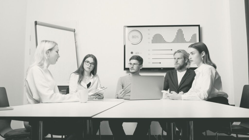 Fünf Personen besprechen sich an einem Konferenztisch, an der Wand ein Bildschirm mit Kennzahlen