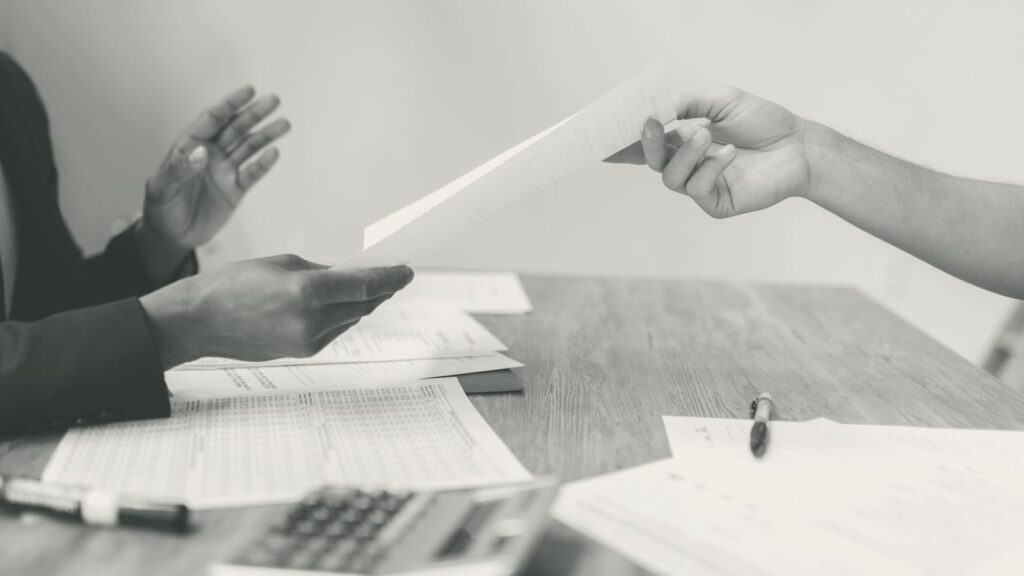 Ein Blatt Papier wird über einen Schreibtisch mit Unterlagen und Taschenrechner von einer Hand in die andere gereicht